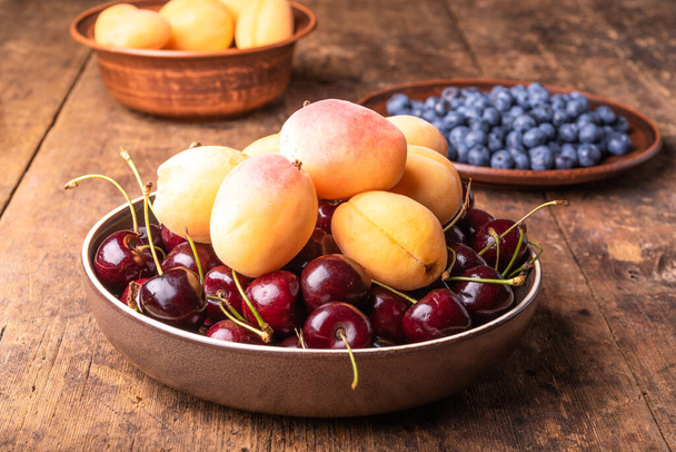 Bagas de damasco maduras saborosas, cerejas e mirtilos em placas em uma mesa de madeira rústica
 - Foto, Imagem