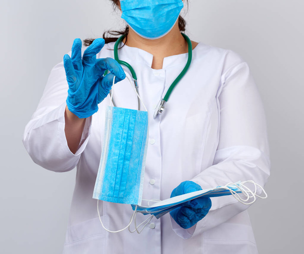 médico fêmea em um casaco branco e máscara detém uma pilha de máscaras protetoras sobre o vírus, médico fica em um fundo branco
 - Foto, Imagem