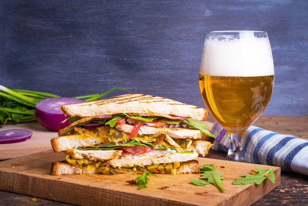 Tasty triangular sandwich made from toasted bread with chicken breast, fried bacon, vegetables, pickles with mustard and arugula closeup and a glass of beer with foam on rustic background, copy space - Photo, Image