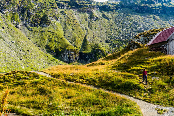 Mies kävelee vuoristopolulla lähellä vuorimajoitusta vuoren ollessa taustalla
 - Valokuva, kuva
