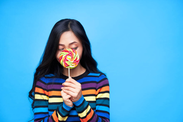 Young woman hide her face behind colorful lollipop isolated on blue background. Emotion cocept, sweet food                       - Photo, image