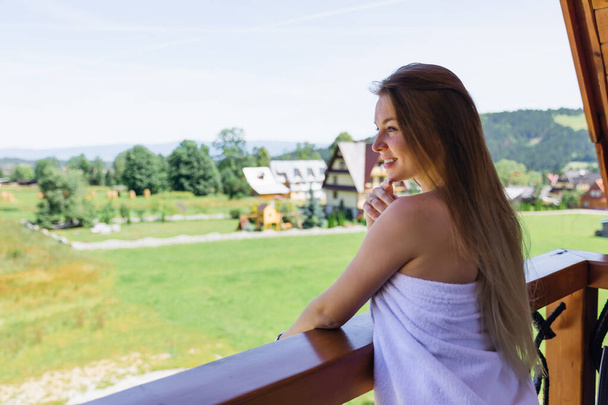 Młoda kobieta stojąca na balkonie w białym ręczniku i podziwiająca widok rano. - Zdjęcie, obraz