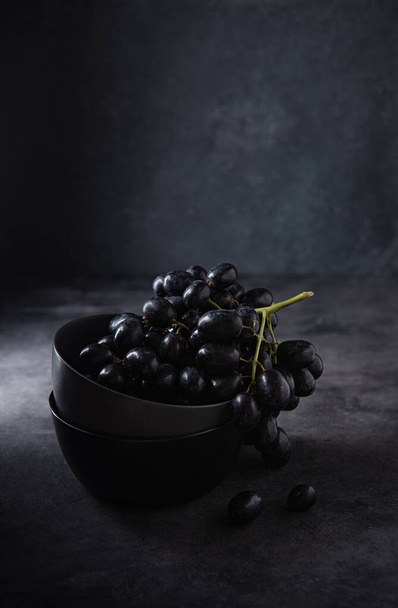 a cluster of juicy black grapes lies in a dark gray bowl on a dark gray table. Photo dark and mood. Front view and copy space - Фото, зображення