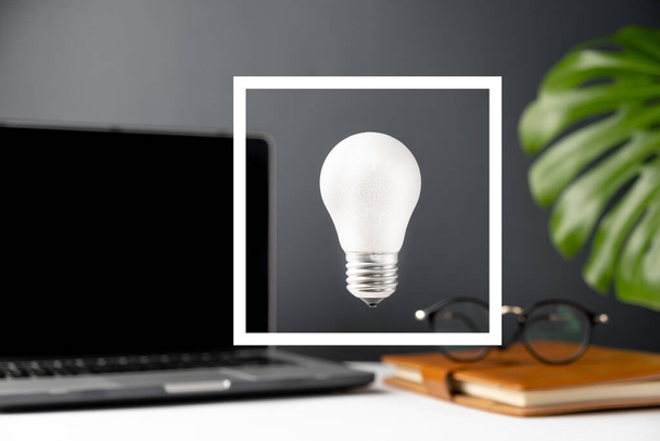 Energy-saving conceptual composition. Home office desk table workspace with a notebook, light bulb, green leaf monstera and eyeglasses on grey background in minimal style. Selective focus - Фото, зображення