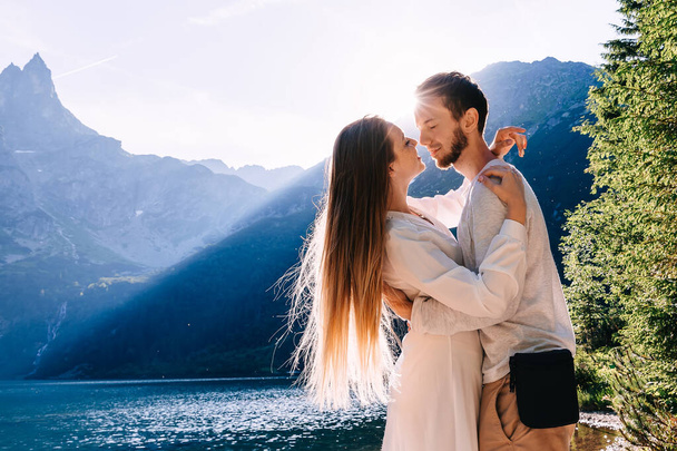 the guy hugs the girl in a white dress for her waist and they close their eyes. sun is shining on the rocky mountains and the lake. - Photo, Image