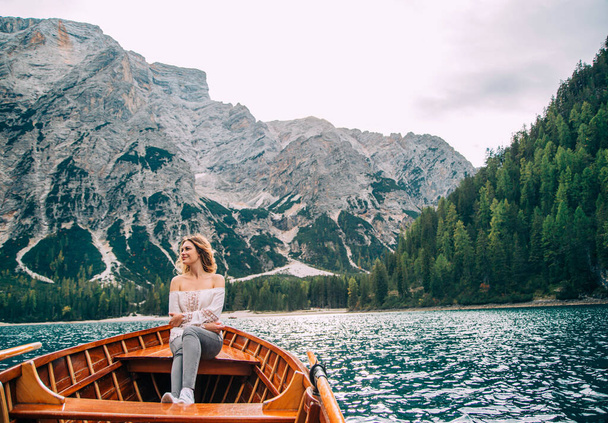Beautiful redhaired girl clothes in white silk sexy blouse long sleeves bare shoulders gray trousers sitting in boat. Tourist enjoy view alpine lake of Braies. Backdrop Italian mountains green forest - Фото, зображення
