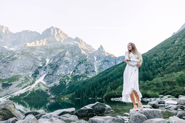 Egy fehér ruhás lány keresztbe tette a karját, és egy nagy kőre állt. sziklás hegyek, tűlevelű erdők és tó. - Fotó, kép
