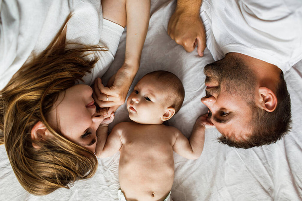 Young attractive happy parents lying in bed upside down with their little baby and looking at her. Wearing white on white blanket background. Clean candid look. - Foto, Imagen