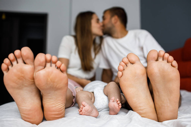 Family feet in focus. Generations living together, lying in bed. Life after parenthood concept. Couple relationships. Parents kissing in background. - Photo, Image