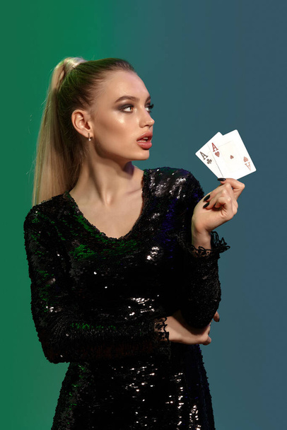 Gorgeous blonde model with ponytail, in jewelry and black sequin dress. She is showing two playing cards, looking aside, posing on colorful background. Gambling, poker, casino. Close-up - Photo, Image