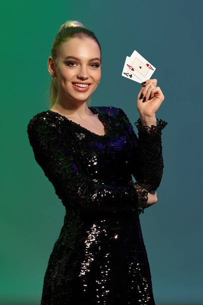 Шикарная блондинка с хвостиком, в ювелирном и черном платье с блестками. Она улыбается, показывая две игральные карты, поддерживая локоть рукой, позируя на красочном фоне. Покер, казино. Крупный план
 - Фото, изображение