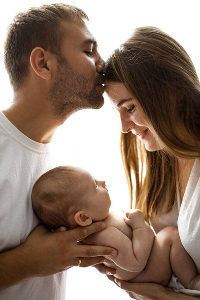 Szczęśliwi rodzice trzymający dziecko i cieszący się cudowną chwilą razem. Mąż całujący żonę w czoło. Podświetlany portret rodzinny na białym tle. - Zdjęcie, obraz