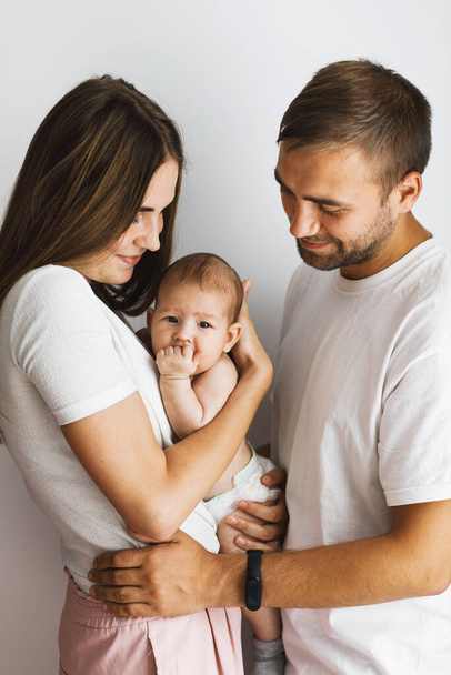 Glückliche junge, gut gelaunte Eltern stehen einander von Angesicht zu Angesicht gegenüber, lächeln und schauen ihr niedliches kleines Baby an. Vertikales Familienporträt. - Foto, Bild