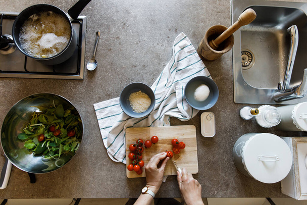 Overhead view of kitchen table during cooking process. Female hands cutting cherry tomatoes on chopping board for salad. Italian cuisine concept. Pasta boiling.  - Photo, image