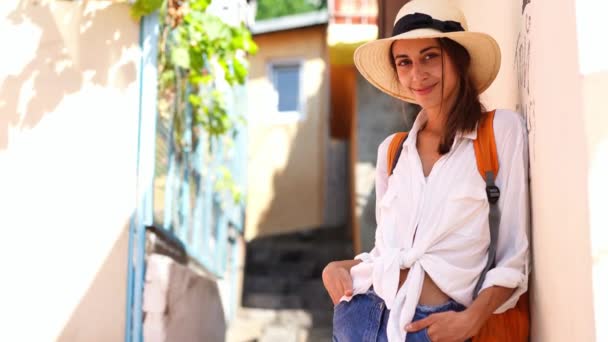 young elegant woman tourist in straw hat, jeans shorts, white shirt and orange backpack enjoying walking narrow street - Felvétel, videó