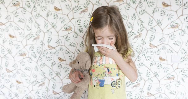 An allergic child sneezes into a handkerchief. Symptoms of influenza and the common cold in primary school children. Girl with a teddy bear caught a cold and sneezes into a napkin - Photo, Image