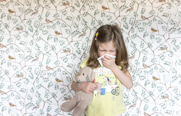 A cute little girl with a teddy bear sneezes into a napkin because she got a viral infection. A sick child with a runny nose coughs and sneezes. Allergic rhinitis in a child. - Photo, Image