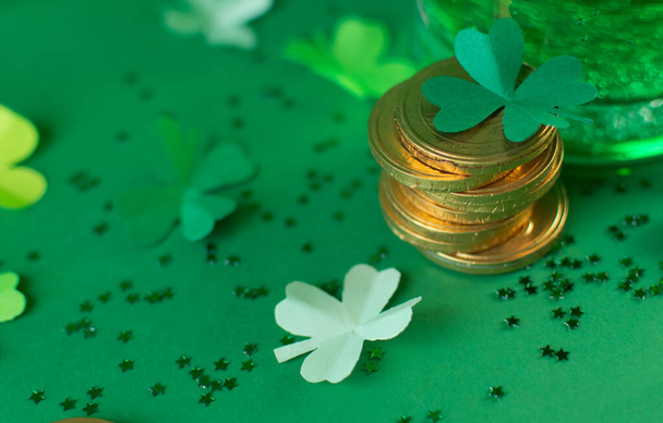 Saint Patricks day backdrop with bear transparent cup with ale, stack of chocolate coins, green four-leafed paper shamrocks on green blurred background with small stars. Lucky concept. Copy space - Foto, afbeelding
