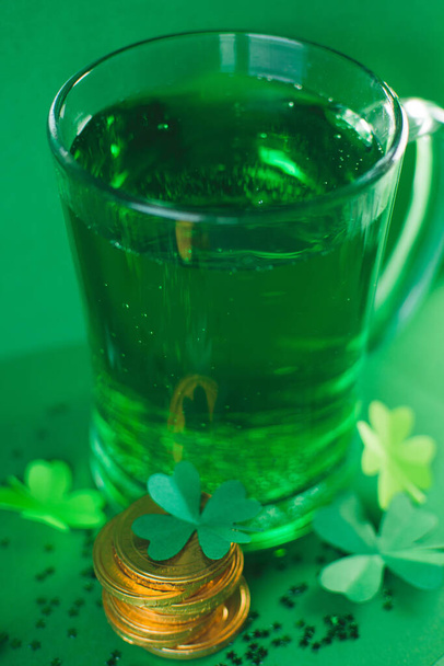 Saint Patricks day backdrop with bear transparent cup with ale, stack of chocolate coins, green four-leafed paper clovers on green blurred background with small stars. Lucky concept. Copy space - Photo, Image