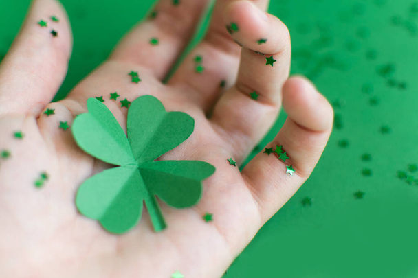Saint Patricks day backdrop with close up green four-leafed paper shamrocks on womans hand sprinkled with small stars on green blurred background. Lucky concept. Copy space. Selective focus - Photo, Image