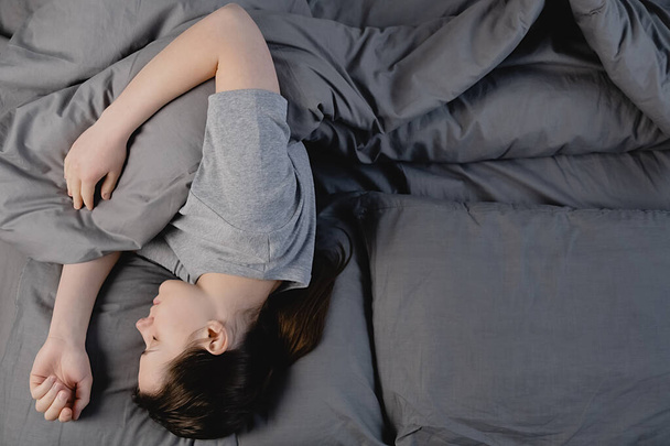Κάτοψη του γαλήνια ήρεμη μελαχρινή γυναίκα κοιμάται σε άνετο κρεβάτι που βρίσκεται σε μαλακό μαξιλάρι ορθοπεδικό στρώμα, ειρηνική νεαρή κοπέλα αναπαύεται καλύπτονται με κουβέρτα σε γκρι σεντόνια στο υπνοδωμάτιο. - Φωτογραφία, εικόνα
