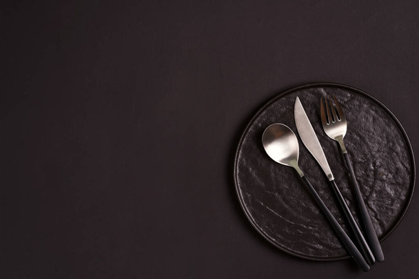 Siyah el yapımı seramik tabakta çatal, kaşık ve bıçak aynı renk arka planda servis ediliyor.. - Fotoğraf, Görsel