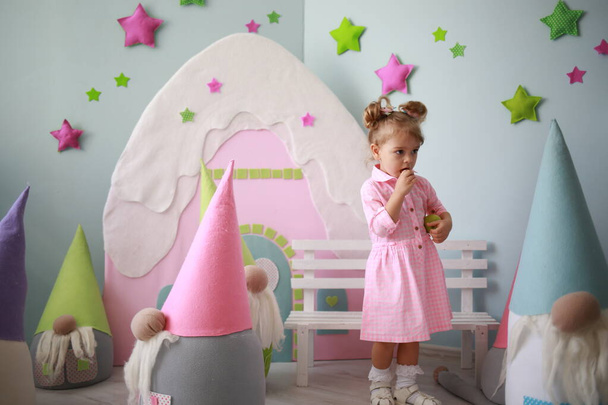 cute caucasian girl in a pink checkered dress has fun in a crowd of large toy gnomes in her bright room - Photo, image