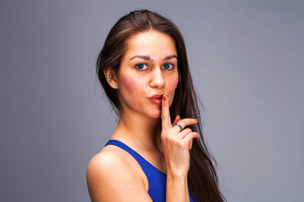 Woman requires silence. Young beautiful brunette model has put forefinger to lips as sign of silence, isolated on gray background - Фото, изображение
