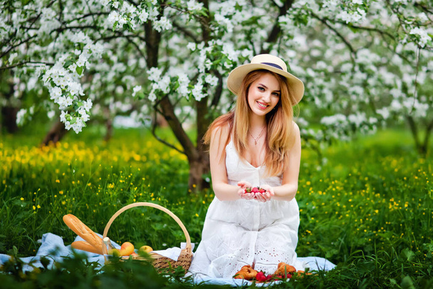 Una hermosa joven sostiene una fresa en sus manos y sonríe. Una encantadora chica blanca con el pelo largo sostiene fresas frescas y jugosas. Concepto de verano y comida saludable
. - Foto, imagen