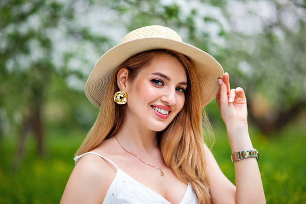 Una donna positiva in cappello e prendisole ride sullo sfondo di un melo in fiore. Una donna ottimista in un ampio sorriso in giardino. Tempo primaverile - Foto, immagini