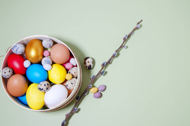 Easter eggs of different sizes: chicken, quail, small candies in the shape of eggs lie in a wooden sieve on a light green background. Nearby lies a willow twig and small eggs. Easter attributes. Hard shadows. Horizontal background - Fotoğraf, Görsel