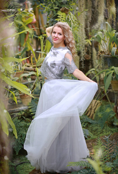 Young beautiful caucasian blonde girl in a glass greenhouse among greenery. Artistic portrait of a girl in a romantic gray dress. - Photo, Image
