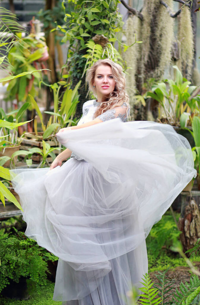 Young beautiful caucasian blonde girl in a glass greenhouse among greenery. Artistic portrait of a girl in a romantic gray dress. - Foto, afbeelding