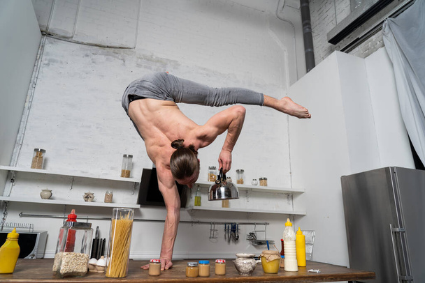 Muscular man stand on one the hand in the kitchen and holding kettle. Concept creativity in the kitchen  - Foto, Imagen