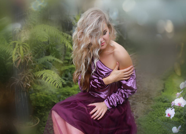 Young beautiful caucasian woman in violet velvet top and lilac tulle skirt, fashion style. In a glass greenhouse among colorful orchid flowers. - Photo, Image