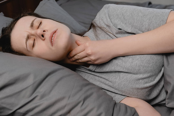 Sick upset girl lying down on bed suffering from throat sore, touching neck, having discomfort or painful feeling, sad female suffering from angina or tonsillitis. Painful swallowing, illness concept - Zdjęcie, obraz