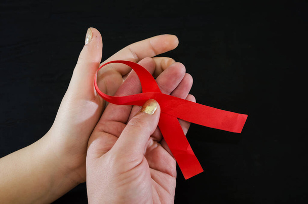 Aids-Konzept. Rotes Band in den Händen auf schwarzem Hintergrund mit Kopierraum. Zwei Hände mit roter Schleife auf schwarz. - Foto, Bild