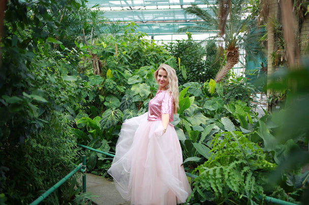 Young beautiful caucasian blond woman in a glass greenhouse among tropical trees. Artistic portrait of a girl in a romantic pink dress. - Photo, Image