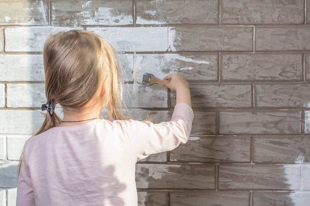 The painter, the plasterer paints a wall. Finishing of facades with brick. Construction and repair work. Brickwork. A little girl will paint a brick wall white. Wall painting, repairs in the house. - Foto, Imagen