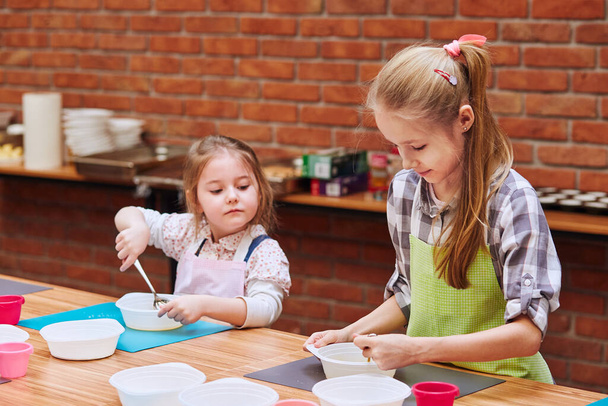 Little girls stirring the yolk with sugar. Kids taking part in baking workshop. Baking classes for children,  aspiring little chefs. Girls learning to cook. Combining and stirring prepared ingredients. Real people, authentic situations - 写真・画像