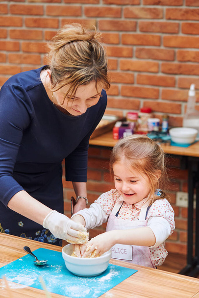 Little girl with her mother's help kneading the dough for baking the cake. Kid taking part in baking workshop. Baking classes for children,  aspiring little chefs. Learning to cook. Combining and stirring prepared ingredients. Real people, authentic  - Fotoğraf, Görsel