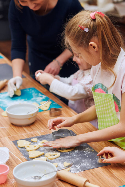 Girls cutting the dough to heart shapes for the cookies. Kids taking part in baking workshop. Baking classes for children, aspiring little chefs. Girls learning to cook. Combining and stirring prepared ingredients. Real people, authentic situations - Fotografie, Obrázek