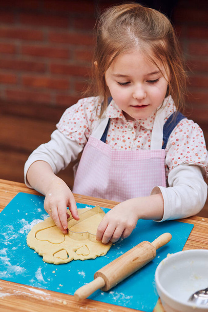 Little girl cutting the dough to heart shapes cookies. Kid taking part in baking workshop. Baking classes for children, aspiring little chefs. Learning to cook. Combining and stirring prepared ingredients. Real people, authentic situations - Photo, Image