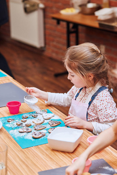 Dívka zdobí své pečené sušenky s barevným posypem a polevou cukru. Dítě, které se účastní pekařské dílny. Hodiny pečení pro děti, ctižádostivé kuchařky. Učím se vařit. Kombinace a míchání připravených ingrediencí - Fotografie, Obrázek