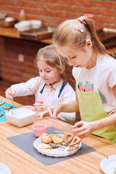 Little girls decorating baked cookies with colorful sprinkle and icing sugar. Kids taking part in baking workshop. Baking classes for children, aspiring little chefs. Learning to cook. Combining and stirring prepared ingredients - Foto, Imagen