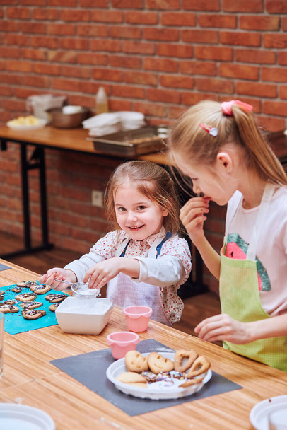 Małe dziewczynki zdobiące ciasteczka z kolorowym posypką i cukrem lukrem. Dzieci biorące udział w warsztatach piekarniczych. Lekcje pieczenia dla dzieci, początkujący mali kucharze. Uczę się gotować. Łączenie i mieszanie przygotowanych składników - Zdjęcie, obraz