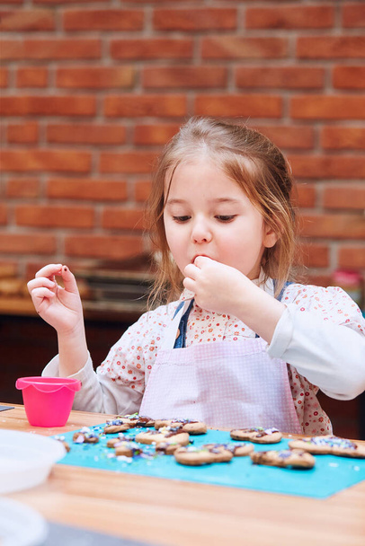 Little girl tasting cookie baked oneself. Decorating her baked cookies with colorful sprinkle and icing sugar. Kid taking part in baking workshop. Baking classes for children, aspiring little chefs. Learning to cook. Combining and stirring ingredient - Foto, Imagem