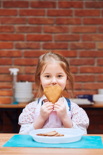 Little girl getting happy out of baked cookie made oneself. Kid taking part in baking workshop. Baking classes for children, aspiring little chefs. Learning to cook. Combining and stirring prepared ingredients. Real people, authentic situations - Photo, image