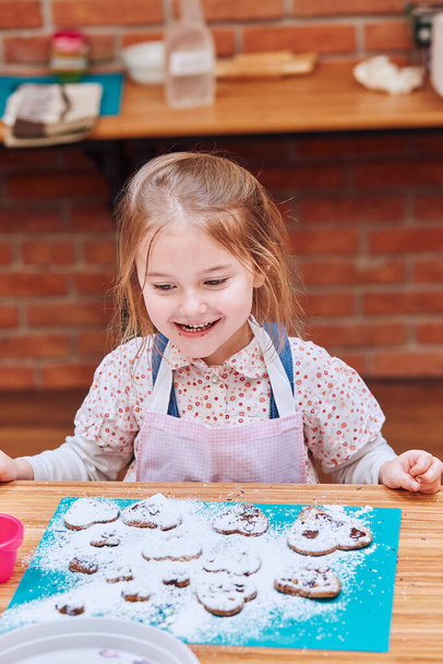 Το κοριτσάκι είναι χαρούμενο εξαιτίας των μπισκότων της. Διακόσμηση μπισκότα με πολύχρωμο πασπαλίζουμε και άχνη ζάχαρη. Το παιδί παίρνει μέρος στο εργαστήριο ψησίματος. Μαθήματα ψησίματος για παιδιά, επίδοξους μικρούς σεφ. Μαθαίνω να μαγειρεύω. Συνδυασμός και ανάδευση συστατικών - Φωτογραφία, εικόνα