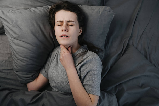 Κάτοψη ανθυγιεινού άρρωστου νεαρή γυναίκα ξαπλωμένη στο κρεβάτι που πάσχουν από πονόλαιμο, αγγίζοντας το λαιμό, έχοντας οδυνηρή αίσθηση, άσχημα λυπημένη γυναίκα που πάσχουν από στηθάγχη ή αμυγδαλίτιδα - Φωτογραφία, εικόνα